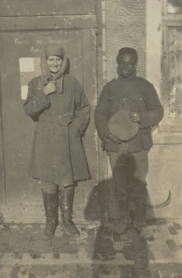Black and white photograph. A pale-skinned woman and dark-skinned man stand against the side of a building. The woman smiles and holds one hand to the scarf draped over her hat and shoulders. The man holds a round object, perhaps a cap, in his hands. The shadow of the photographer falls over the bottom half of the man’s body.