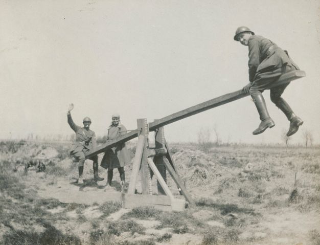 Black and white photograph. Two soldiers use a wooden structure as a seesaw. Both look at us and the lower one, farther away, waves. A woman smiles next to him. Scrubby plants grow on the flat, otherwise barren, land.